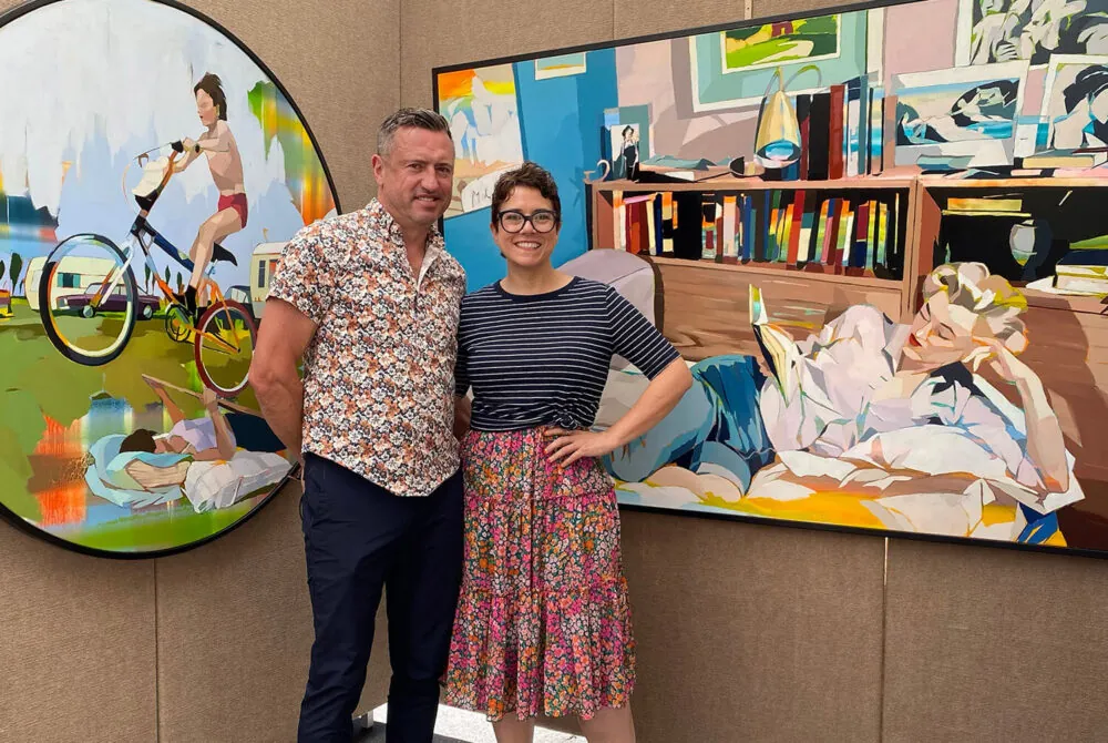 Photo of Signe & Genna Grushovenko smiling in an artist booth with vibrant paintings